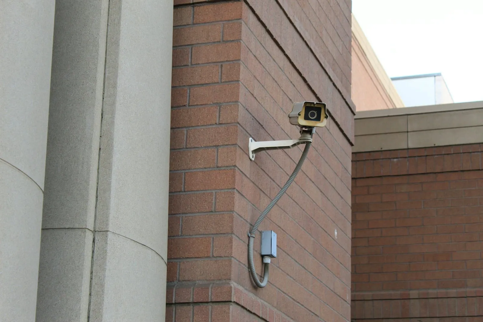 A security camera mounted on a red brick wall next to a concrete pillar